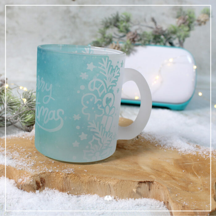 Eine satinierte / gefrostete Tasse die mit einer weihnachtlichen Banderole sublimiert wurde. Sie ist in hellblau, so dass es wie Eis aussieht. Darauf steht der Schriftzug "Merry Christmas". Im Hintergrund steht ein Cricut Joy, mit dem die Banderole erstellt wurde.