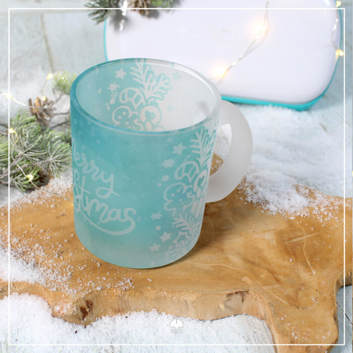 Eine satinierte / gefrostete Tasse die mit einer weihnachtlichen Banderole sublimiert wurde. Sie ist in hellblau, so dass es wie Eis aussieht. Darauf steht der Schriftzug "Merry Christmas". Im Hintergrund steht ein Cricut Joy, mit dem die Banderole erstellt wurde.