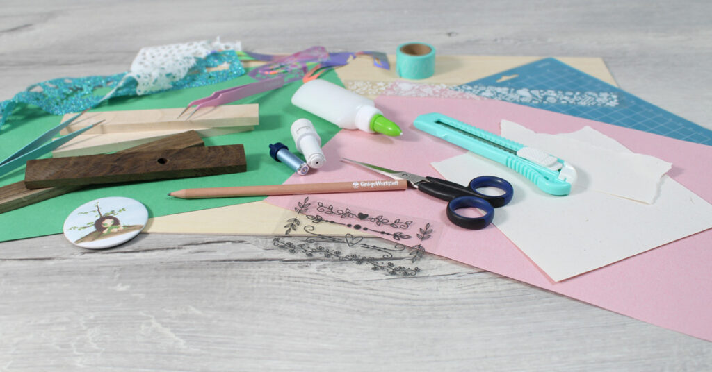 Auf einem Tisch liegen verschiedene Materialien aus dem Kreativbereich. Unter anderem Plottermesser, Holz, Papiere, Scheren, Kleber, Pinzetten, Schneidematten und mehr.