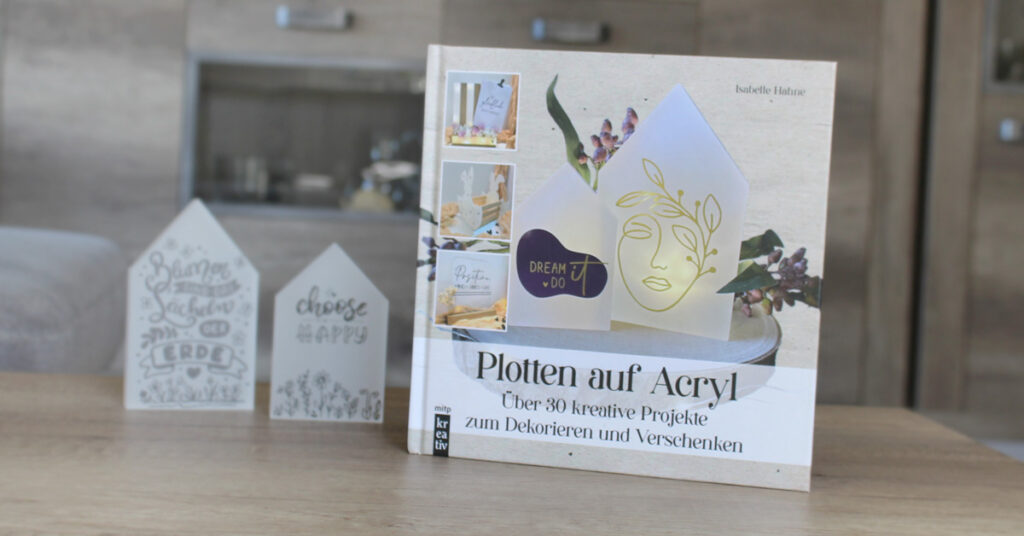 Beitragsbild zum Blogartikel "Buchvorstellung: Plotten auf Acryl". Zu sehen sind das Buch, sowie zwei kleine Acrylhäuschen. Diese wurden mit RubOns verziert.