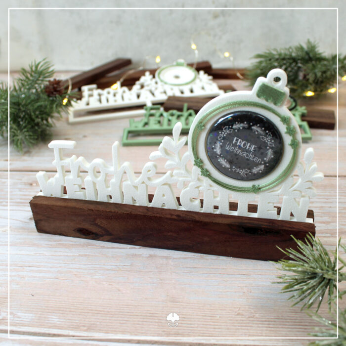 Abgebildet ist die 38er Version der 3D Klötzchenschrift "Frohe Weihnachten" - 3D Druckdatei in weiß. Es ist ein schwarzer Button eingesetzt und der Einschieber wurde zwischen zwei dunkle Klötzchen geklebt.