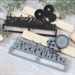 Abgebildet sind zwei Varianten der 3D Klötzchenschrift "Herzlichen Glückwunsch" - 3D Druckdatei, welche mit Buttons kombiniert werden können. Die Version für den 25er Button wurde einfarbig aus schwarzem Filament auf einer Holoplatte gedruckt. Für den 32er Button wurde der Einschieber grau und die Details schwarz gedruckt.