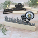 Abgebildet sind zwei Varianten der 3D Klötzchenschrift "Herzlichen Glückwunsch" - 3D Druckdatei, welche mit Buttons kombiniert werden können. Im Vordergrund ist eine Version für die 32er Buttons, welche in grau und schwarz gedruckt wurde. Der Einschieber befindet sich zwischen hellen Klötzchen aus Bergahorn.