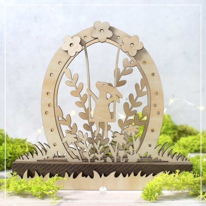 Ein Mini-Lichterbogen aus drei Ebenen mit Klötzchen kombiniert. Das Motiv zeigt einen Hasen auf einer Schaukel in einer Ei-Silhouette im Gras. Die Motive wurden aus Holz mit dem Laser geschnitten.