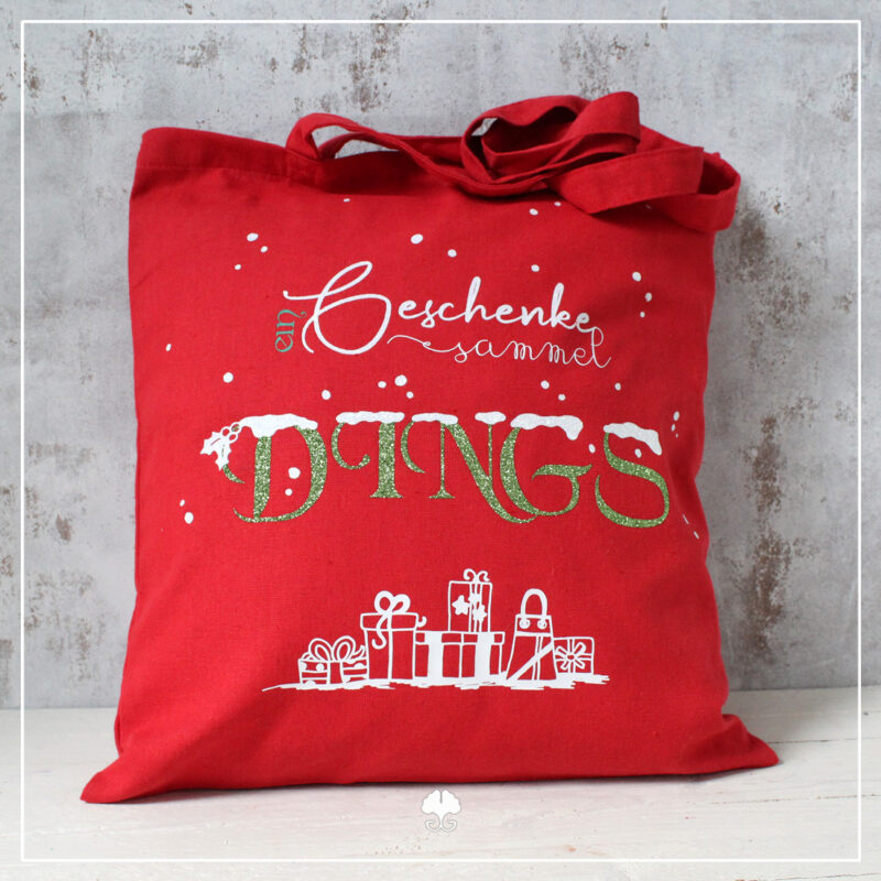 Ein roter Beutel auf dem das Wortspiel "Geschenkedings" aufgeplottet wurde. Für den Schriftzug wurden Glitzerfolien in grün und weiß genutzt. Die Geschenke am unteren Rand sind aus einer weißen Folie geplottet.
