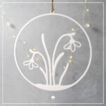 Ein weißer, 3D-gedruckter Schneeglöckchen-Kranz, filigran gedruckt, hängend an einer Jutekordel vor einem grauen Hintergrund mit stimmungsvollen, bokeh-artigen LED-Lichtern.