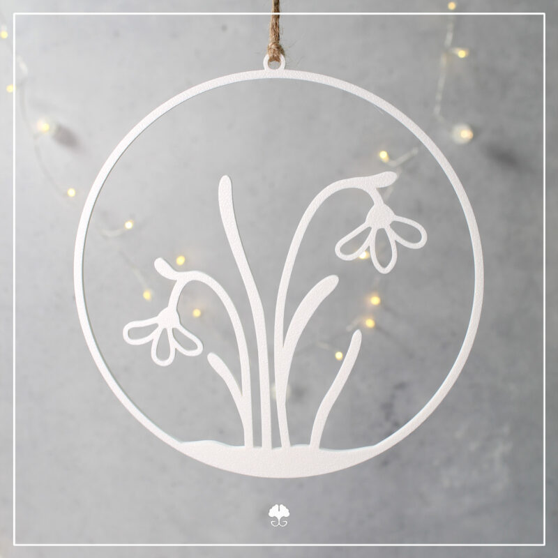 Ein weißer, 3D-gedruckter Schneeglöckchen-Kranz, filigran gedruckt, hängend an einer Jutekordel vor einem grauen Hintergrund mit stimmungsvollen, bokeh-artigen LED-Lichtern.