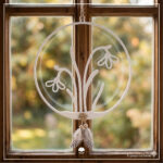 Ein weißer, hängender 3D-Druck-Kranz mit Schneeglöckchen, verziert mit Jutekordel, Holzperlen, einer kleinen Holzblume und gepunkteten Federn. Er hängt vor einem hellen Fenster mit Aussicht auf einen Garten im weichen Fokus.