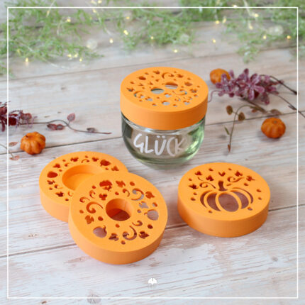 Vier orangefarbene 3D-gedruckte Blumenretter- und Kerzenhalter-Aufsätze mit Kürbismotiv neben einem Glück-Glas.