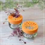 Glück-Gläser mit aufgeschraubten herbstlichen Blumenrettern im Kürbis-Design auf hellem Holz.