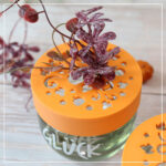 Detailaufnahme eines 3D-gedruckten Blumenretters mit herbstlichen Pflanzen auf einem Glück-Glas.